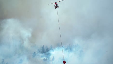 Helicopter-with-water-bucket,-flying-over-smoking-wildfire-forests,-in-Western-USA