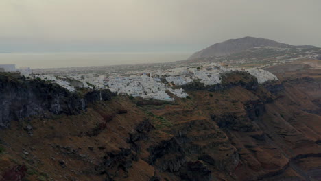 Aerial:-Slow-panning-drone-shot-of-Fira-in-Santorini,-Greece-during-cloudy-sunrise