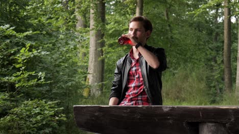 Young-adult-man-having-break-during-hiking-in-forest-to-drink-by-wooden-table