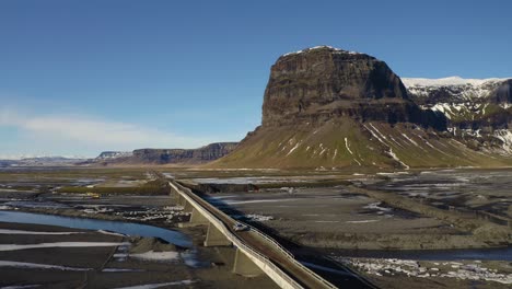 Vista-Aérea-Del-Coche-Cruzando-El-Puente-Lomagnupur-En-Islandia-Con-Una-Enorme-Montaña-En-El-Fondo---Hermoso-Día-Soleado-Con-Cielo-Azul-En-La-Isla