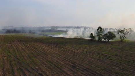 Steady-aerial-footage-of-a-tilled-farmland-and-then-burning-while-white-smoke-blown-to-the-right,-Grassland-Burning,-Pak-Pli,-Nakhon-Nayok,-Thailand