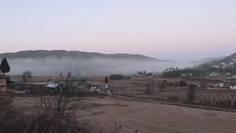 Pequeño-Pueblo-Remoto-En-Las-Faldas-De-Las-Montañas-Con-Niebla-Y-Cielo-Plano-Por-La-Mañana-Desde-Un-ángulo-Plano