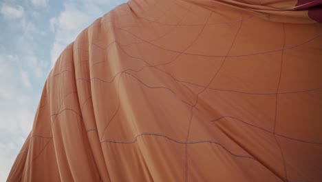 Orange-color-hot-air-balloon-deflating-on-ground-against-blue-sky,-handheld-view