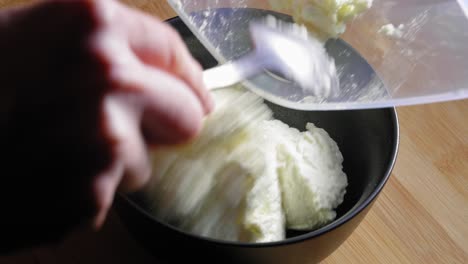 A-close-up-shot-of-a-black-mixing-bowl-on-a-wooden-kitchen-table,-a-chef-carefully-preparing-white-low-fat-cheese-by-transferring-it-from-a-storage-container-and-placing-it-into-the-bowl