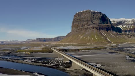 Filmische-Luftaufnahme-Der-Idyllischen-Isländischen-Landschaft-Mit-Flussdelta,-Autoüberquerungsbrücke-Und-Riesigem-Berg-Lomagnupur-Im-Hintergrund-Bei-Sonnenlicht-In-Island