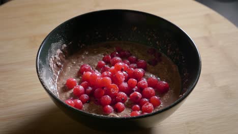A-close-up-shot-tracking-in-onto-a-low-calorie-protein-pudding-topped-with-sweet-tasty-red-currants,-a-delicious-healthy-dessert-served-in-a-black-bowl-on-a-wooden-kitchen-table