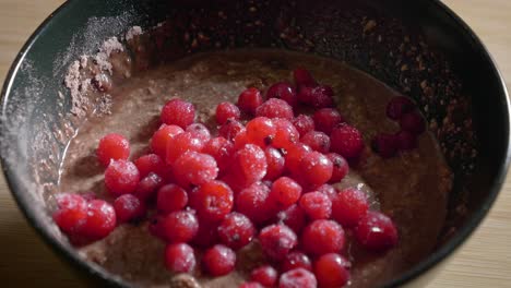A-close-up-shot-of-a-black-dessert-bowl-filled-with-a-mixture-of-oatmeal,-protein-powder-and-low-fat-cheese,-as-tasty-frozen-red-currants-are-added-to-the-dish-creating-a-low-calorie-protein-pudding