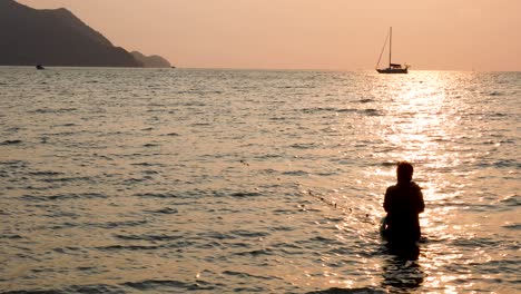 Eine-Frau,-Die-Gegen-Einen-Schönen-Sonnenuntergang-Und-Eine-Yacht-Im-Hintergrund-Am-Horizont-In-Bangsaray-In-Der-Nähe-Von-Pattaya,-Thailand,-Fischt