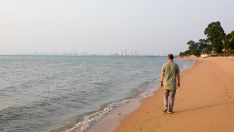 Male-Tourist-Walking-Away-Along-the-Beach-of-Bangsaray-in-Pattaya,-Thailand-with-City-View-in-the-Distance