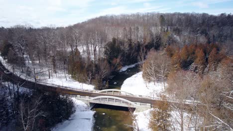 Winter-Aerial-of-Car-driving-over-bridge-through-dense-forest-landscape-river-covered-in-fresh-snow