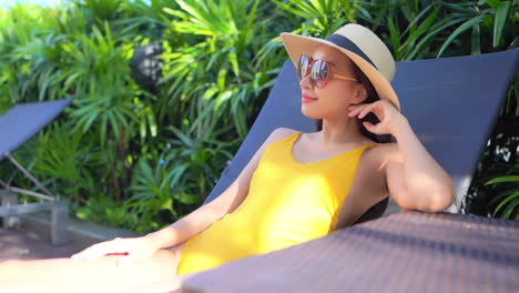 Close-up-of-an-attractive-woman-in-a-yellow-bathing-suit,-sunglasses,-and-straw-sun-hat-relaxes-poolside-in-a-lounge-chair