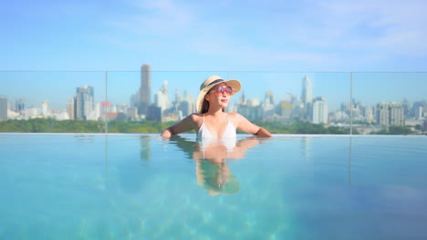 With-the-city-skyline-behind-her,-an-attractive-woman-lounges-in-the-water-along-the-edge-of-a-resort-pool
