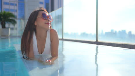 Close-up-of-a-pretty-woman-lying-on-her-stomach-in-the-rooftop-swimming-pool-as-she-looks-out-over-a-modern-city-skyline