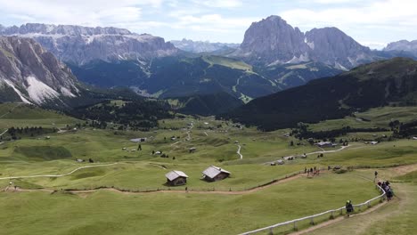 Val-Gardena-Valley-to-the-Seceda-Mountain-Peak-at-South-Tyrol,-Italian-Alps,-Dolomites,-Italy---Aerial-Drone-View-of-Hikers,-Green-Valley,-Cabins-and-Mountains
