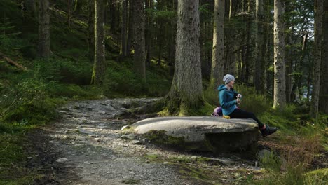 A-woman-sits-on-a-rock-eating-her-lunch-and-drinking-tea-from-a-flask-while-hiking-in-the-forest,-looking-out-across-a-Scottish-Loch-and-hillside-in-the-sun