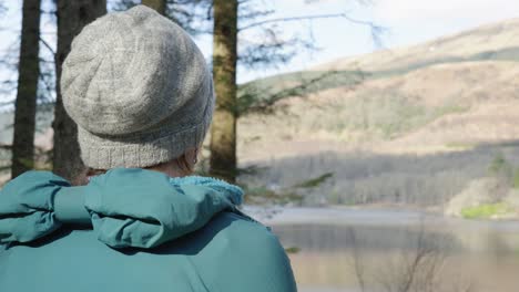 A-camera-looks-over-the-shoulder-of-a-woman-eating-her-lunch-while-out-for-a-walk-in-the-forest,-looking-out-across-a-Scottish-Loch-and-hillside-in-the-sun