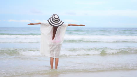 Back-to-the-camera-a-young-fit-woman-in-a-flopping-sun-hat-and-white-cover-up-welcomes-the-surf-onto-the-beach