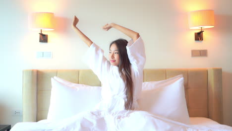 Young-attractive-Asian-woman-sitting-in-bed-wearing-white-robe-stretches-with-arms-above-head-to-wake-up-in-morning