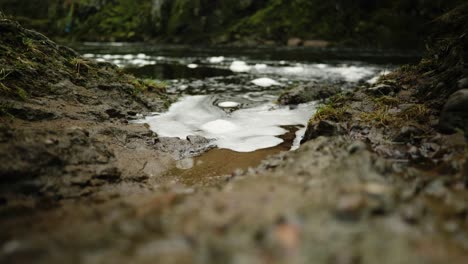 Schaum-Und-Blasen-Schwimmen-Auf-Der-Wasseroberfläche-Des-North-Esk-River-In-Schottland,-Der-Langsam-Gegen-Die-Ränder-Eines-Schlammigen-Flussufers-Schwappt,-Während-Im-Hintergrund-Ein-Schnell-Fließender-Fluss-Vorbeifließt