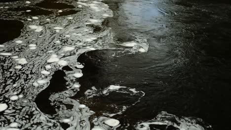 Time-lapse-of-swirling-bubbles-of-white-foam-floating-on-the-surface-of-dark-water-in-a-Scottish-river-in-a-constantly-changing-pattern-where-the-faster-current-meets-a-still-section-of-water