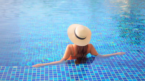 Woman-in-orange-swimsuit-and-straw-cap-relaxing-in-clear-warm-water-of-luxury-resort-swimming-pool