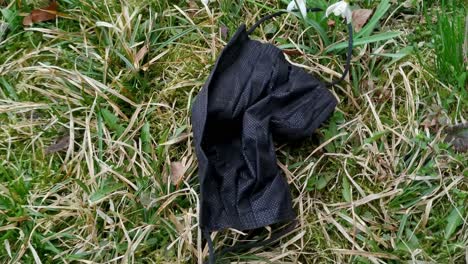 Closeup-of-black-surgical-face-mask-on-ground-grass,-pandemic-pollution,-handheld,-day