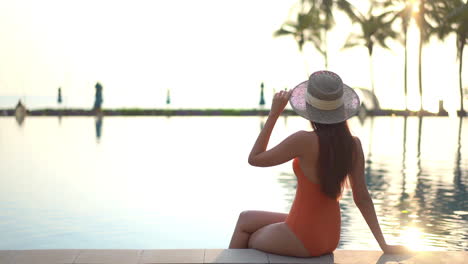 Back-to-the-camera,-a-woman-sitting-on-the-edge-of-a-pool-reaches-up-to-adjust-her-sun-hat