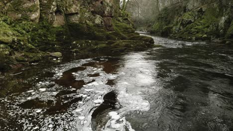 Swirling-bubbles-of-white-foam-float-slowly-on-the-surface-a-Scottish-river-in-a-hypnotic,-constantly-changing-pattern-of-organic-shapes-as-a-fast-river-current-flows-through-a-green,-mossy-gorge