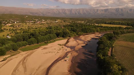 People-relaxing-on-sandy-river-during-dry-season-with-mountains-in-background-at-sunset,-Cordoba-in-Argentina
