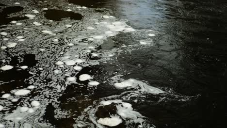 Swirling-bubbles-of-white-foam-slowly-float-on-the-surface-of-dark,-black-water-in-a-Scottish-river-in-a-hypnotic,-constantly-changing-pattern-where-the-faster-current-meets-a-still-section-of-water