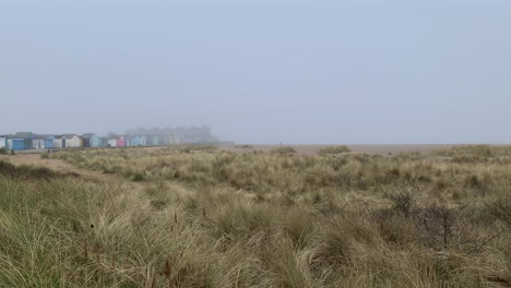 Beach-huts-on-a-misty-winter-coastline-and-grassy-sand-dunes-beneath-a-foggy-grey-sky