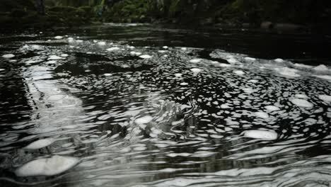 Swirling-bubbles-of-white-foam-floats-on-the-surface-of-dark,-black-water-in-a-Scottish-river-in-a-hypnotic,-constantly-changing-pattern-as-a-shallow-depth-of-field-highlights-the-centre-of-the-frame