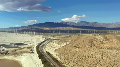 Drone-flies-over-train-tracks-towards-windmill-farm-in-Palm-Springs,-San-Bernardino-Mountains-in-distance