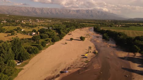 Luftaufnahme-Eines-Sandigen-Ausgetrockneten-Flusses-Mit-Parkenden-Autos-Während-Der-Ferien-In-Cordoba,-Argentinien---Idyllische-Landschaftsfelder-Und-Bergkette-Im-Hintergrund