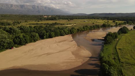 Dry-river-with-verdant-banks-and-mountains-in-background,-Cordoba,-Argentina