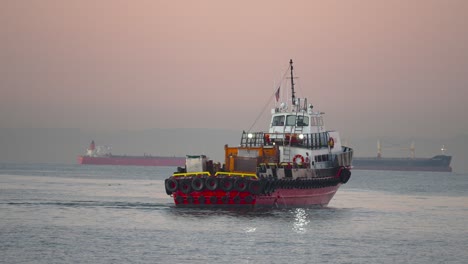 Ein-Schlepper-Sticht-Bei-Einem-Farbenprächtigen-Sonnenaufgang-Mit-Containerschiffen-Im-Hintergrund-In-See