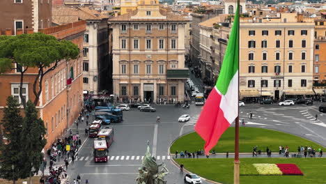 Aerial-shot-of-traffic-on-roundabout-in-old-town-of-Rome---In-foreground-italian-flag---View-from-Altar-of-Fatherland