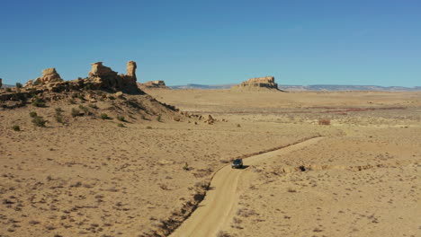 Aerial-as-car-drives-across-open-desert-with-rock-formations-in-distance