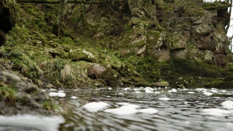 Schaumblasen,-Die-Auf-Der-Wasseroberfläche-Des-North-Esk-River-In-Schottland-Schwimmen,-Wirbeln-Langsam-Auf-Die-Kamera-Zu-Und-In-Eine-Flache-Fokusebene-Mit-Einer-Moosbedeckten-Klippe-Im-Hintergrund