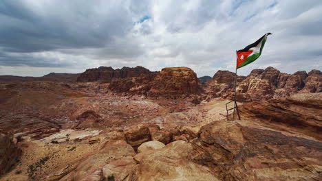 Cinemagraph-seamless-video-loop-of-the-Jordanian-flag-in-Petra,-Jordan,-moving-in-the-wind-at-a-bedouin-camp-with-vibrant-colors-and-a-view-of-the-desert-rocks-with-tombs-carved-into-sandstone-rock