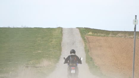 A-motorcyclist-riding-along-a-dusty-gravel-road-between-two-meadows-in-Germany