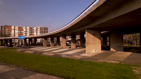 Slow-aerial-sideways-underneath-large-concrete-overpass-with-thick-columns-of-highway-intersection-in-urban-surrounding