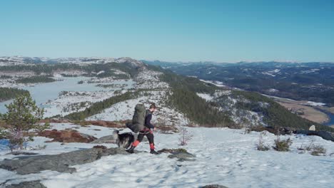 Un-Hombre-Mochilero-Con-Su-Perro-Caminando-En-La-Ladera-De-La-Montaña-ártica-En-Un-Día-Soleado-De-Invierno-En-Noruega
