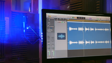 An-audio-waveform-on-a-computer-monitor-in-a-recording-studio-with-microphone-visible-in-the-vocal-booth-behind