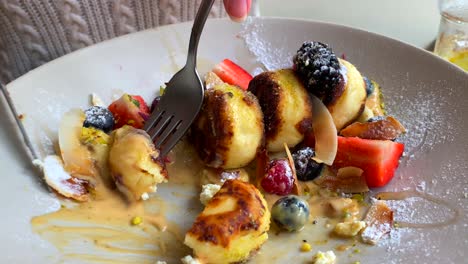 Eating-delicious-syrniki-with-forest-berries,-banana-and-coconut-shavings-in-cashew-cream-and-honey,-tasty-mini-breakfast-cheese-pancakes,-traditional-ukrainian-russian-dish,-4K-shot