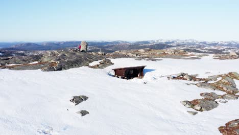 Dos-Turistas-Excursionistas-En-La-Cima-De-Una-Montaña-Cubierta-De-Nieve-Con-Una-Pequeña-Cabaña-De-Invierno-De-Madera