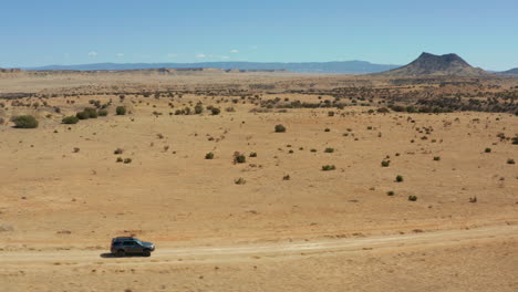 Aerial-of-car-driving-down-dirt-road-with-vast-desert-landscape-in-background