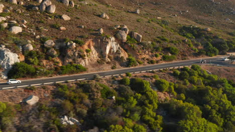 White-Car-Driving-At-Victoria-Road-Passing-By-Mountain-Hills-With-Boulders-In-South-Africa