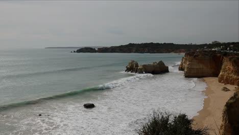Wide-slow-motion-view-from-cliffs-above-Praia-do-Vale-do-Olival,-Portugal-on-an-overcast-afternoon-with-cloudy-horizon-over-water-and-waves-crashing-against-huge-rocks