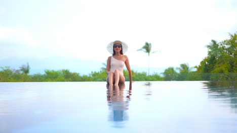 With-the-ocean-at-her-back,-an-attractive-woman-sits-in-the-shallow-water-on-the-edge-of-a-resort-pool-and-adjusts-her-sunglasses-as-she-looks-around